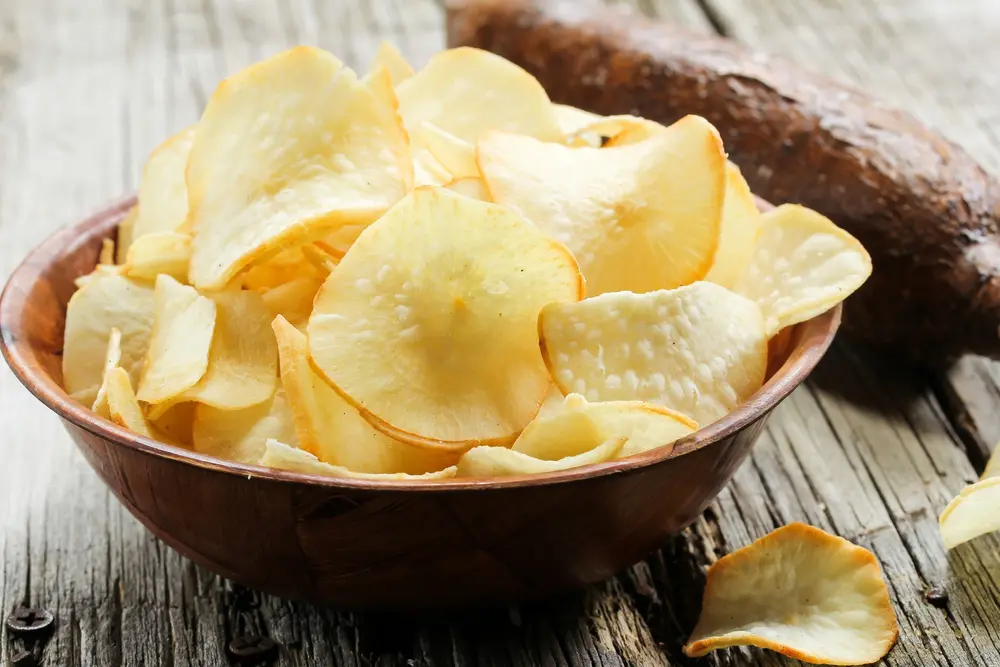 Crispy golden cassava chips served in a rustic wooden bowl next to raw manioc root tubers